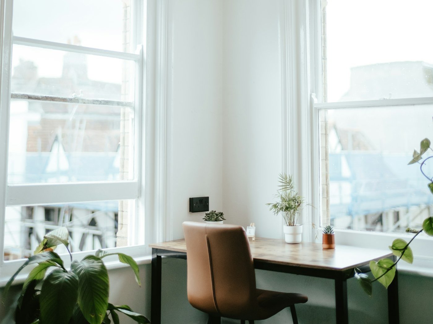 Small wooden desk and chair in a bright corner between two tall windows, with plants